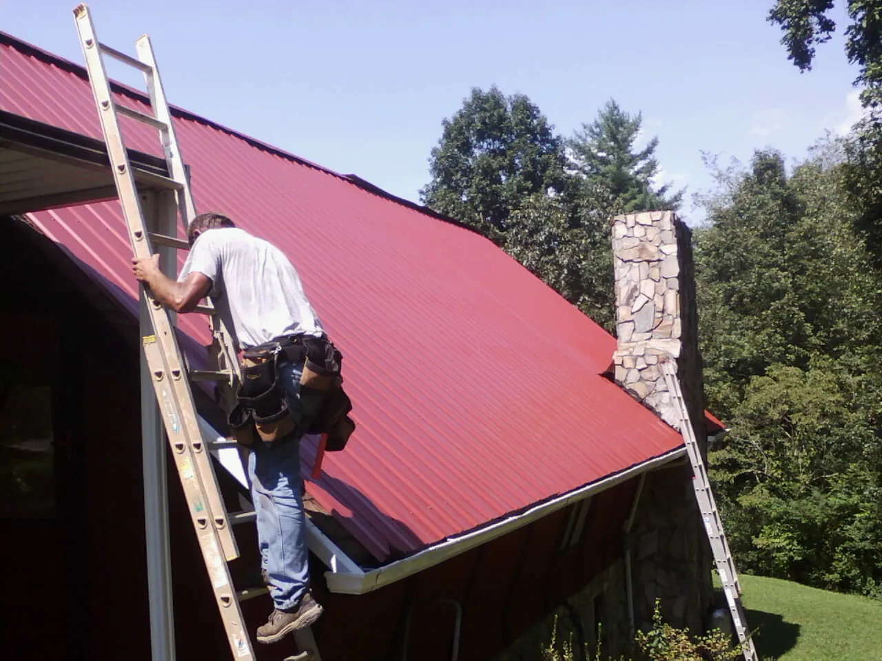 Roofer on a ladder beside a red metal roof and stone chimney — Soffit & Fascia Repair by Roofing Brothers in Gurnee